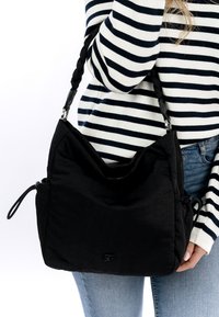 Zwarte stoffen schoudertas met een gestructureerde afwerking, voorzien van gevlochten banden en een verstelbare trekkoord. Logo prominent weergegeven aan de voorkant.