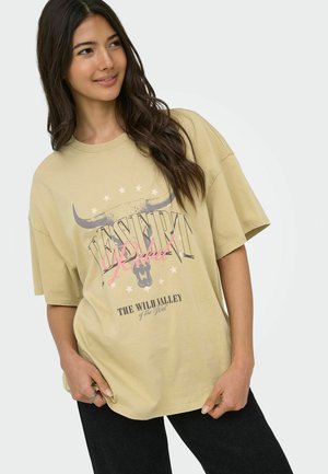Ung kvinde med langt mørkt hår iført en beige oversized T-shirt med teksten "DESERT Rebel" og en grafik af et okseskal.