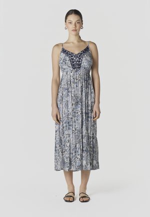 Noor naine seisab, kandes sinise-valge lillemustrilise midi kleiti, millel on õhukesed rihmad, ning musti sandaale, taust on ühtlane.