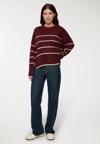 Młoda kobieta ubrana w bordowy sweter w białe paski, niebieskie dżinsy i białe trampki, stoi z jedną ręką na biodrze.
