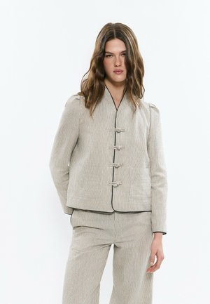 Femme portant une veste beige à fines rayures et un pantalon assorti avec des boutons noués, debout devant un fond blanc.