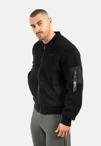 Hombre con cabello corto que lleva una chaqueta negra con textura y cremallera, con un bolsillo con cremallera y una etiqueta en la manga izquierda, y pantalones grises, contra un fondo liso.