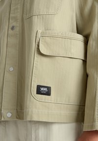Chaqueta en espiga de color beige claro con un bolsillo en el pecho izquierdo, cierre de botón a presión plateado y un parche con el logo negro de Vans. Textura suave, diseño casual.