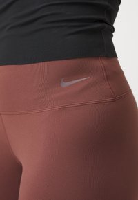 Nike leggins v rjavo-rdeči tkanini z gladko teksturo. Značilnosti vključujejo logo na pasu in podrobnosti šivanja.