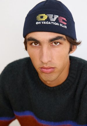 Jeune homme aux yeux marron portant un bonnet bleu marine brodé "OVC On Vacation Club" et un pull sombre à rayures bleu et rouille.