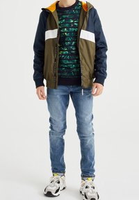 Cortavientos verde y marino con franja blanca, capucha naranja; superpuesto sobre un suéter de patrón de hojas verde oscuro; jeans azules y zapatillas blancas.