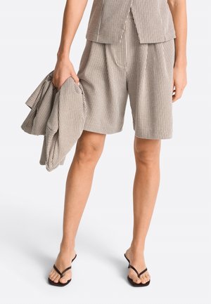 Person trägt beige-weiß gestreifte Shorts und passendes Weste, hält Jacke, mit schwarzen, riemchen Sandalen an nackten Füßen.