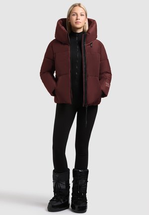 Maroon puffer jacket with a large hood, zip front, and side pockets. Worn over a black ribbed outfit and complemented by black insulated boots.