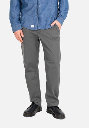 Man wearing grey pants, black lace-up shoes, and a blue long-sleeve button-up shirt, standing with one hand in pocket.