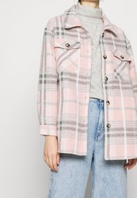 Veste à carreaux rose et grise avec une fermeture à boutons sur le devant, deux poches poitrine et un col, fabriquée en tissu texturé.