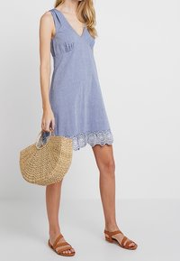 Blaues Leinen-Kleid ohne Ärmel mit V-Ausschnitt, gerafften Details und einem mit Spitze verzierten Saum. Kombiniert mit einer geflochtenen Strohtasche und braunen Sandalen.
