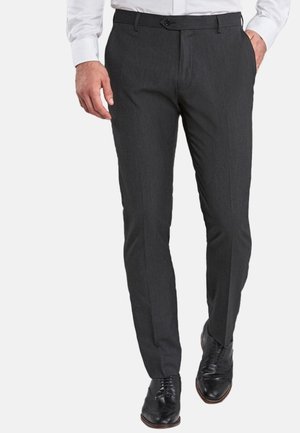 Homme portant un pantalon habillé gris foncé slim, des chaussures en cuir noir et une chemise blanche à manches longues, marchant avec une main dans la poche.