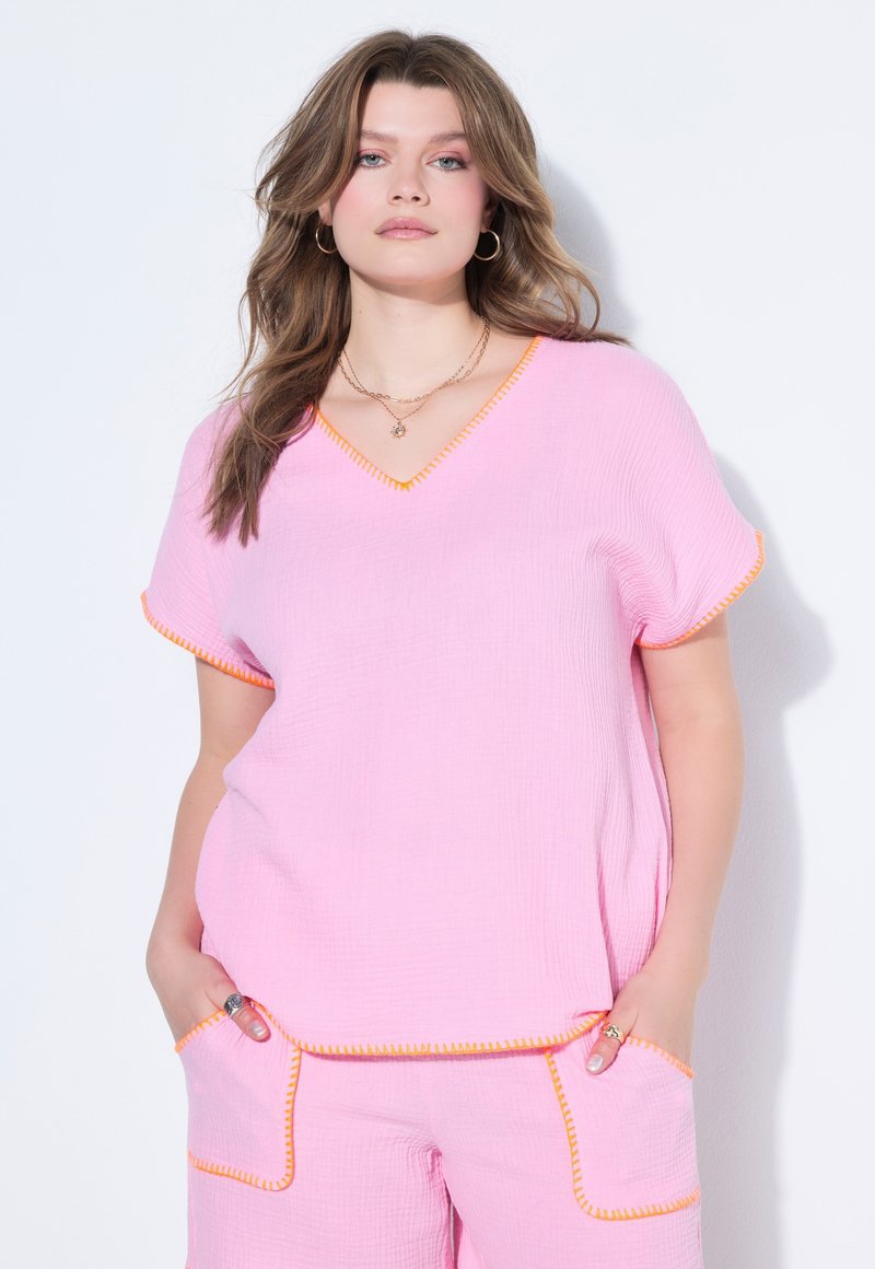 Ung kvinde iført lyserød tekstureret kortærmet top og matchende shorts med orange syningskant, hænderne i lommerne, står mod en hvid baggrund.