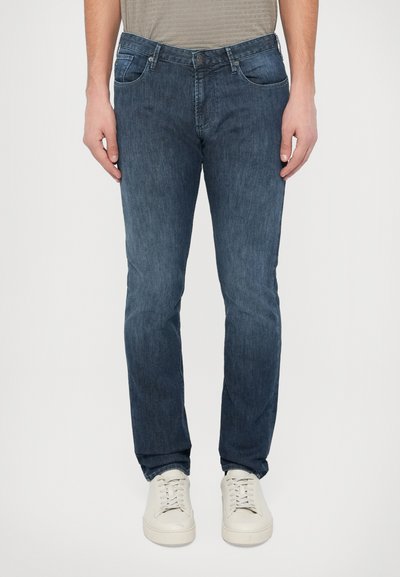 Denim jeans in dark blue with a faded wash, five-pocket design, and straight-leg fit. Paired with white sneakers for contrast.