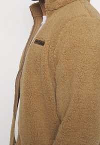 Brun fleecejacka med hög krage, dragkedja framtill och en mjuk strukturerad yta. Har en liten logotyp på bröstet.