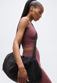 Νεαρή γυναίκα με αθλητική περιβολή μωβ χρώματος, κρατώντας μια μαύρη τσάντα γυμναστηρίου στον ώμο της, μπροστά σε ένα απλό φωτεινό φόντο.