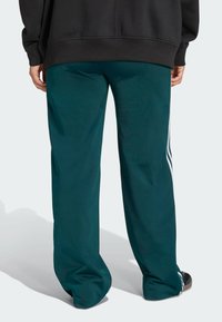 Teal track pants made of smooth fabric, featuring white side stripes, a relaxed fit, and a wide leg design; worn with a black sweatshirt.