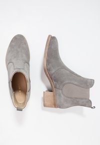 Botines de ante gris con puntera redondeada, paneles laterales elásticos y tacón grueso de madera. Cuentan con una lengüeta en la parte trasera para facilitar su puesta.