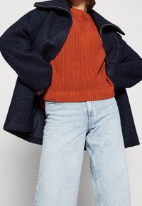 Navy villasekoituksesta valmistettu takki, jossa on vetoketjullinen kaulus, yhdistetty oranssiin ribbiknitteiseen neuleeseen ja vaaleansinisiin korkeavyötäröisiin farkkuihin. Kangasrakenteen yksityiskohdat näkyvissä.