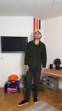 Tamnozeleno korduroy jakna s dva džepa na grudima i dugmadima, uparena s crnim trapericama i crnim slip-on cipelama, u modernom enterijeru.