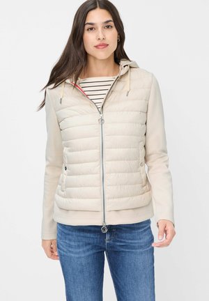 Frau trägt eine beige gesteppte Jacke mit Kapuze und ein gestreiftes Hemd, kombiniert mit blauen Jeans, steht vor einem weißen Hintergrund.