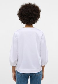 Weiße Bluse mit Ballonärmeln, runder Halsauschnitt und glatter Textur. Getragen mit blauen Jeans, von hinten betrachtet.