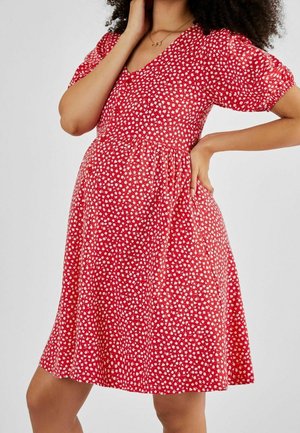 Kobieta ubrana w czerwoną sukienkę w kwiaty z krótkimi rękawami, stojąca z jedną ręką na biodrze, a drugą przy twarzy.