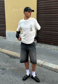 Bílá košile s dlouhými rukávy a černými rukávy, grafický potisk a text 2025; šedé džínové šortky; černé boty s perforovanou texturou; bílé ponožky.