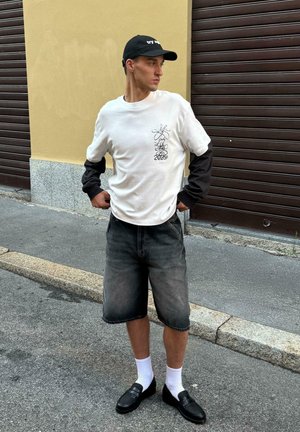 Hvid langærmet skjorte med sorte ærmer, grafisk print og 2025 tekst; grå denim shorts; sorte sko med perforeret tekstur; hvide sokker.