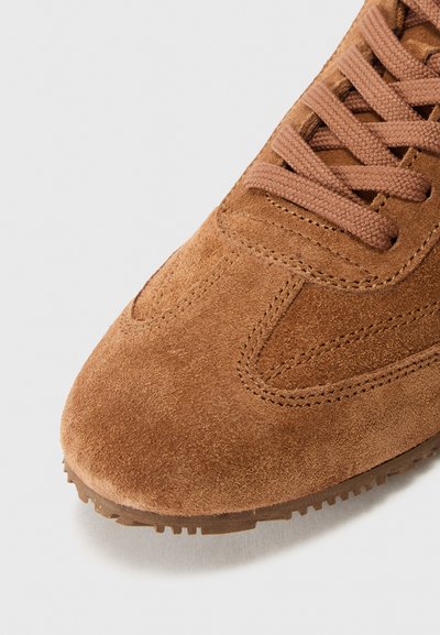 Sneaker en suede marron avec un bout arrondi, une surface texturée et des lacets plats. La semelle extérieure dispose d'un motif pour une meilleure adhérence.