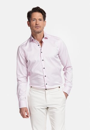 Lichtroze overhemd met lange mouwen, een puntkraag en zwarte knopen. Model draagt witte broek en staat tegen een effen achtergrond.