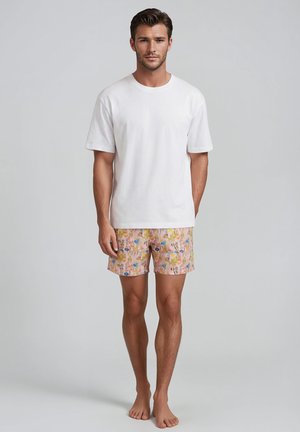 Man staat op blote voeten, draagt een effen wit T-shirt met korte mouwen en roze korte broek met bloemenpatroon tegen een grijze achtergrond.