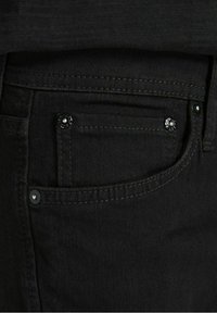 Jeans de mezclilla negra con una textura suave, adornados con acentos metálicos en el bolsillo delantero y costuras en contraste a lo largo de los bordes.