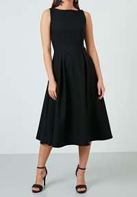 Černé bezrukávové šaty s přiléhavým živůtkem a rozšířenou sukní, vyrobené z hladké tkaniny. V kombinaci s černými lodičkami na kotníkové pásky.