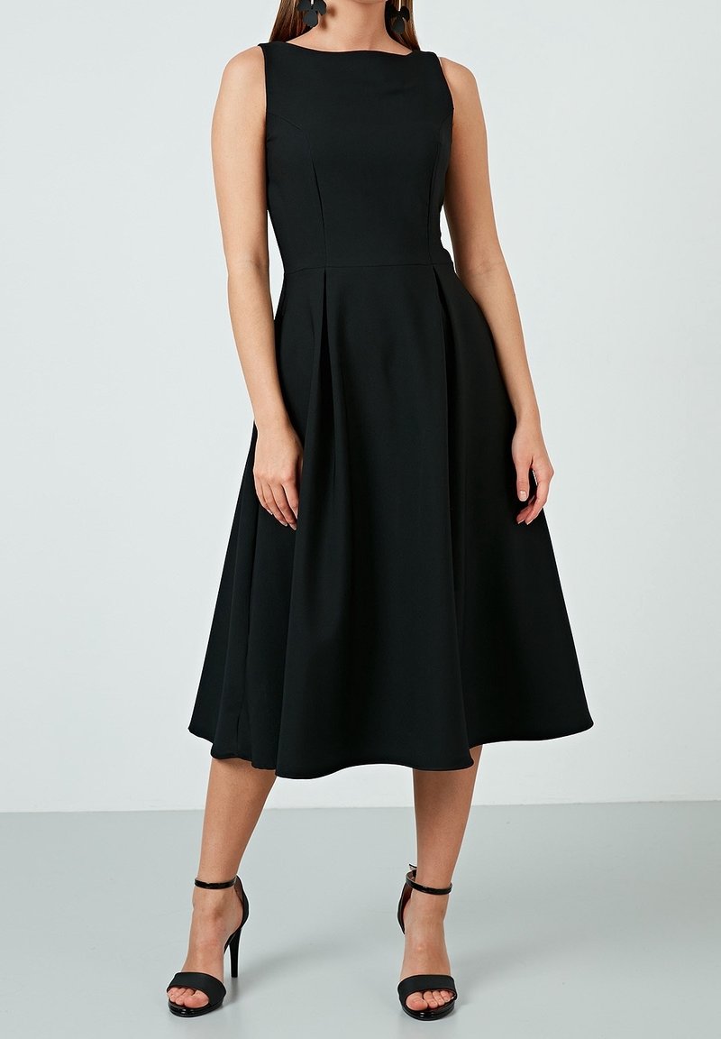 Černé bezrukávové šaty s přiléhavým živůtkem a rozšířenou sukní, vyrobené z hladké tkaniny. V kombinaci s černými lodičkami na kotníkové pásky.