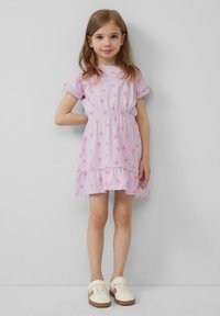 Robe rose avec motif de fraises, taille élastique, manches courtes avec liens, ourlet festonné, associée à des baskets crème sur un fond uni.