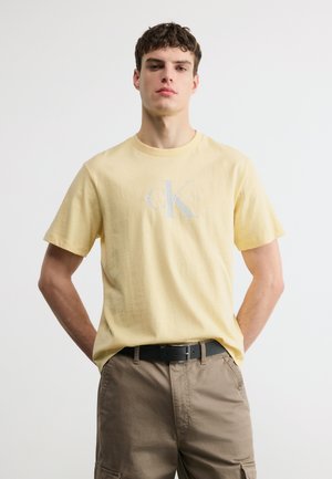 Νέος άνδρας που φοράει ένα ανοιχτό κίτρινο t-shirt Calvin Klein, φορεμένο μέσα σε μπεζ cargo παντελόνι με μαύρη ζώνη, στέκεται μπροστά σε λευκό φόντο.