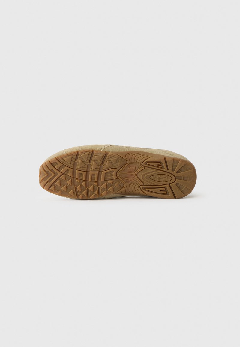 Light tan sneaker outsole featuring a zigzag tread pattern, textured rubber for grip, and branded detailing near the heel.