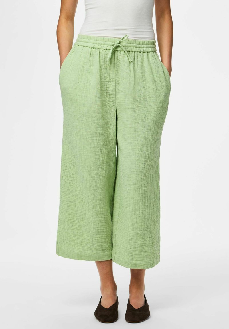 Femme portant un pantalon large court vert clair avec cordon à la taille et poches, associé à des chaussures noires sans lacets.