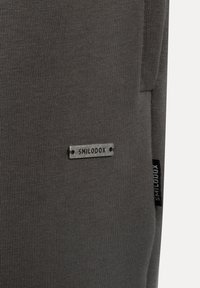 Grå stofstruktur med lodret ribbing. Har en sølvmetalplade med "SMILODOX" graveret på. En lille sort label er synlig.