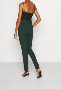 Green tailored trousers with a fitted design, high waist, and back pockets, paired with a black camisole and black high-heeled sandals.