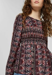 Langs mouw blouse met een elastische taille in een veelkleurig geometrisch patroon. De stof toont tinten van rood, zwart en wit met textuurdetails.
