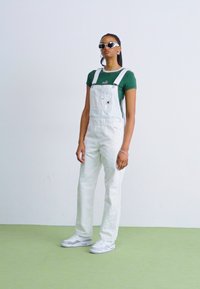 Salopette en denim blanc avec des bretelles ajustables, portée sur un t-shirt vert. Le mannequin porte des lunettes de soleil et des baskets blanches sur un sol vert clair.