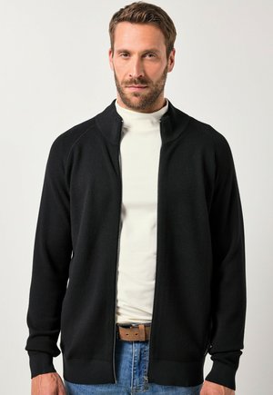 Zwarte ritscardigan met geribbelde kraag en boorden, gemaakt van zacht breimateriaal. Gekleed over een witte turtleneck, gecombineerd met blauwe spijkerbroek.