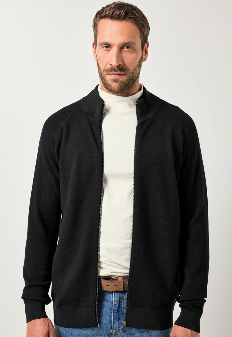 Cardigan zip-up nero con colletto e polsini a costine, realizzato in materiale a maglia morbido. Indossato sopra un dolcevita bianco, abbinato a un paio di jeans blu.