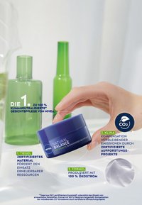 Blue jar labeled "NIVEA Natural Balance," featuring a smooth surface. Green glass bottles in the background. Hand reaching for the jar.