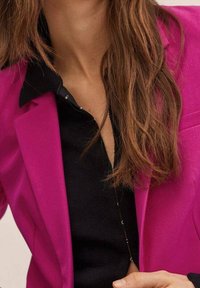 Jasnoróżowa marynarka na czarnej koszuli, z wyciętym kołnierzem i guzikami, wykonana z gładkiej tkaniny o prostym kroju.