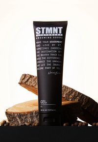 Schwarze Tube Haar-Gel mit der Aufschrift "STMNT" in weißer Schrift, ausgestattet mit einem Klappverschluss. Vor einem strukturierten Holzhintergrund platziert.