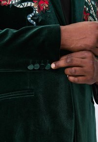 Emerald green velvet blazer with embroidered floral and snake design. Four fabric buttons on cuff, subtle sheen, smooth texture.