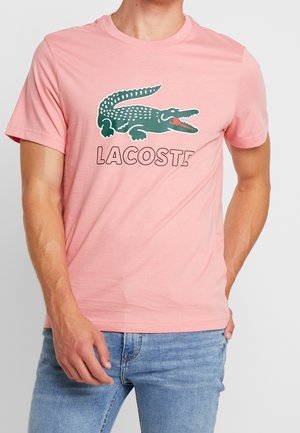 Homme portant un t-shirt rose avec un logo crocodile vert et un texte "LACOSTE", associé à un jean bleu clair.
